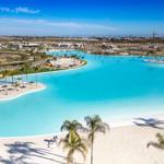 Large pool with sandy areas and palm trees under blue sky.