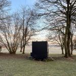 Black trailer on grass field between bare trees with view of field.