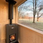 Sauna with wooden walls and wood-burning stove, view of field and trees.
