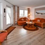 Wohnzimmer mit zwei orangefarbenen Sofas, runden Teppich und Holztisch. Fenster mit Vorhängen.