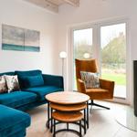 Ein Wohnzimmer mit blauem Sofa, orangefarbenem Sessel und Kaminofen. Große Fenster und Wandbilder.