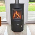 Ein Kaminofen mit brennendem Holz steht vor einer Glasfront. Ein brauner Sessel ist links im Bild.