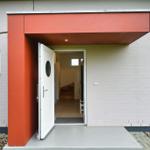 Haus mit rotem Vordach und weißer Eingangstür. Durchgang ins Innere mit Holztreppe.