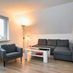 Wohnzimmer mit grauem Sofa, Sessel und Tisch. Fenster und Wandbilder sind sichtbar.