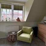 Ein Zimmer mit grünem Sessel, Holzschrank und Fenster mit Jalousien.