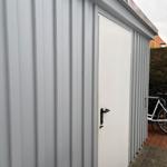 Graue Metallgarage mit weißer Tür und schwarzer Klinke. Fahrrad steht daneben.