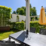 Terrasse mit Tisch, Stühlen und gelber Sonnenschirm. Grünfläche und Bäume im Hintergrund.