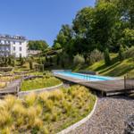 Pool mit Holzterrasse in grüner Umgebung. Im Hintergrund steht ein weißes Gebäude.