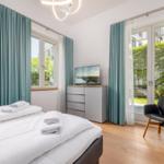 Schlafzimmer mit Bett, TV, Stuhl und Fenstern mit Blick auf Grün.