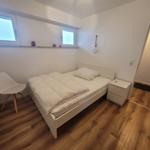 Schlafzimmer mit weißem Bett, Nachttisch und Stuhl. Holzboden und Fenster.