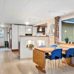 Dining area with wooden table and blue chairs, kitchen with white cabinets and window with colorful curtains.