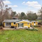 Yellow vacation house with terrace, garden, and trees. White bench and table are outside.