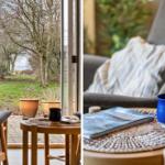 A gray chair with cushions and table with blue mug and book. View of garden and woods.