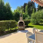 Terrace with grill, table and chairs. Green garden and trees in the background.
