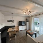 Living area with dining space, TV, and balcony access. Wood floor and white furniture.
