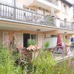 Haus mit Balkon und Garten am Wasser. Rote Sonnenschirme und Pflanzen sind sichtbar.