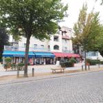 Strandbuchhandlung und Geschäfte am gepflasterten Weg mit Bäumen und einem Sitzbänkchen.