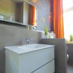 Bad mit Waschbecken, Spiegel und Fenster mit orangefarbenen Vorhängen.