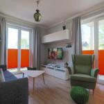 Wohnzimmer mit Sofa, Sessel und Fernseher. Große Fenster mit orangefarbenen Jalousien.