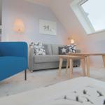 Ein Wohnzimmer mit grauem Sofa, blauem Sessel und Holztisch unter einem Dachfenster.