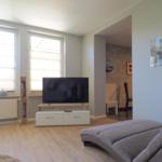 Wohnzimmer mit Sofa, Fernseher und Fenstern. Blick in die Küche.