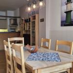 Essbereich mit Holztisch und Stühlen, Küchenbereich im Hintergrund, Wandbild eines Leuchtturms.