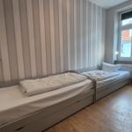 Zimmer mit zwei Einzelbetten, gestreifter Wand und Holzboden. Fenster mit Blick auf Gebäude.