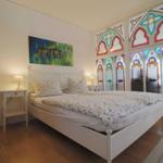 Ein Schlafzimmer mit weißem Bett, Nachttischen und bunten Glasfenstern.