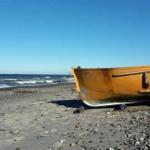 Ein orangefarbener Bootsrumpf steht auf einem steinigen Strand vor dem Meer.