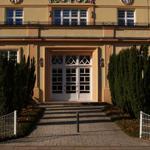 Eingangsbereich mit weißen Türen und Zierhecken vor einem hellen Gebäude.