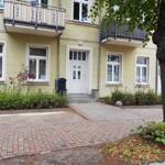 Haus mit Eingang, Balkonen und gepflastertem Weg. Grünflächen und Baum im Vorgarten.