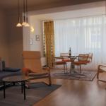 Wohn- und Essbereich mit Sofa, Sessel und Tisch. Helle Fenster mit weißen Vorhängen.
