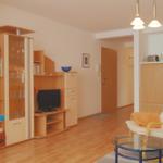 Wohnzimmer mit Holzmöbeln, Fernseher und Sessel. Küche im Hintergrund.