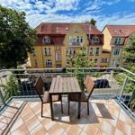 Terrasse mit Tisch und zwei Stühlen, Blick auf Wohnhäuser und Bäume unter blauem Himmel.