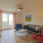 Wohnzimmer mit Sofa, Stühlen und TV. Balkon mit Liegestühlen. Wandbild und Deckenventilator.
