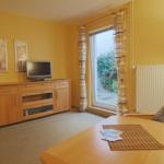 Wohnzimmer mit Fernseher, Tisch und Sessel. Fenster mit Vorhängen. Wand mit Bildern.