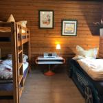 Room with bunk bed and car bed, wooden walls, table with lamp and pictures.