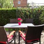 Terrasse mit Tisch und Stühlen, eine Blumenvase und grüne Hecke im Hintergrund.
