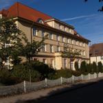 Ein mehrstöckiges Gebäude mit rotem Dach und weißen Fenstern. Vor dem Haus steht ein weißer Zaun.
