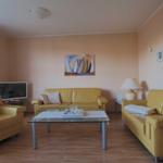 Wohnzimmer mit zwei gelben Sofas, Glas-Tisch und Wandbild mit Segelbooten.