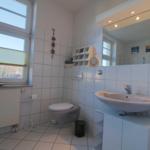 Badezimmer mit Toilette, Waschbecken und Fenster. Weiße Fliesen und Spiegel.