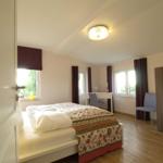 Schlafzimmer mit Bett, Schreibtisch, Stühlen und Fenstern mit Vorhängen.