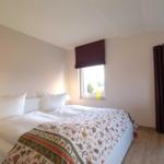 Schlafzimmer mit Bett, Fenster und Vorhängen. Holzboden und weißer Wandschrank.