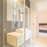 Badezimmer mit Dusche und Waschbecken. Im Hintergrund Schlafzimmer mit Bett.