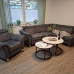 Wohnzimmer mit grauem Sofa, Sitzgelegenheit und Tischen. Fenster mit Jalousien und Pflanzen.