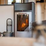 Ein Kaminofen mit brennendem Holz steht im Wohnbereich. Im Hintergrund sind Küchenschränke zu sehen.