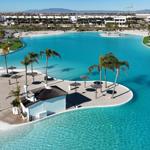 Large pool with island, palms, and umbrellas. Background: residential area and mountains.