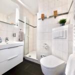 Badezimmer mit Dusche, Waschbecken und Toilette. Weiße Fliesen und dunkler Boden.