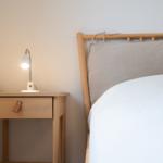 Ein Bett mit Holzgestell und weißer Bettwäsche. Neben dem Bett steht ein Nachttisch mit einer Lampe.