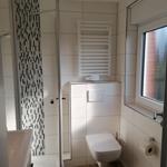 Badezimmer mit Dusche, Toilette und Fenster. Weiße Fliesen und Holzfußboden.